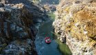 Aerial shot of a red raft floating down the Rogue River through a tight gorge with the right side in the sun and the left side of the gorge in the shade.