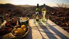 Three metal wine glasses in front of two bottles of white wine next to a tray of chips and other light snacks overlooking the African desert at sunset