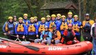 Group of rafters on land wearing blue splash jackets, yellow helmets, and orange life jackets smiling before they get on the river.